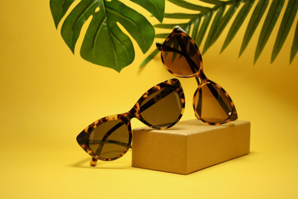 occhiali da sole poggiati su un blocco di legno su sfondo giallo e piante verdi